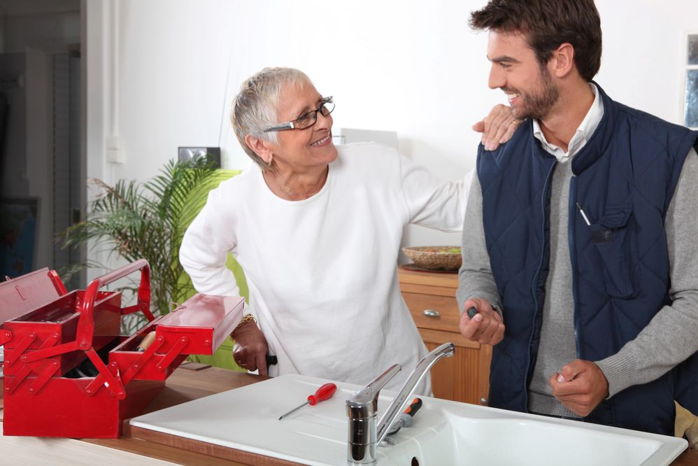A plumber helping an elderly homeowner with kitchen sink repairs inside her home.